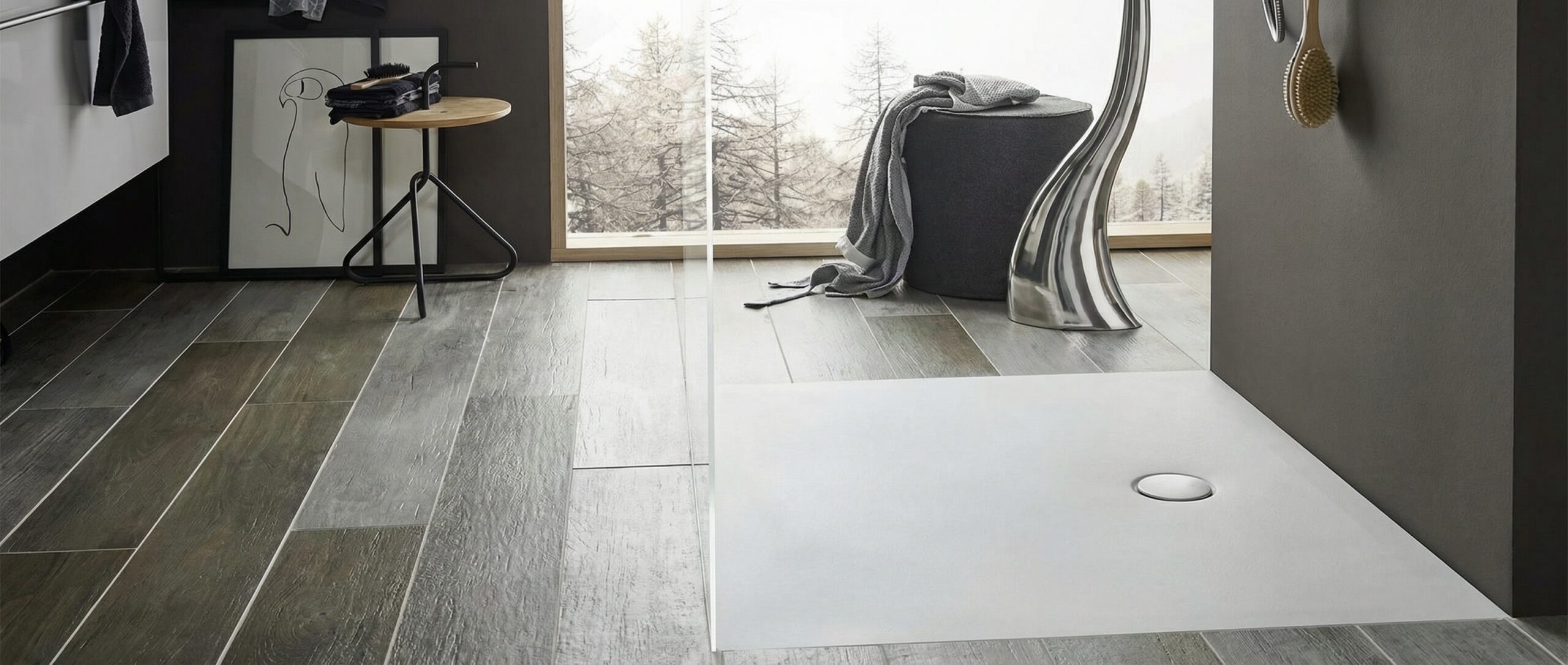 Modernes Badezimmer mit dunklen Holzoptik-Fliesen und einer ebenerdigen, weissen Duschtasse mit rundem Abfluss. Eine durchsichtige Glaswand trennt den Duschbereich. Ein grosses Fenster im Hintergrund bietet Blick auf verschneite Bäume. Im Raum stehen ein dunkler Pouf mit einem grauen Handtuch, eine hohe silberne Dekoskulptur und ein kleiner Holztisch mit schwarzen Handtüchern und einer Bürste. Ein minimalistisches gerahmtes Bild lehnt an der Wand.