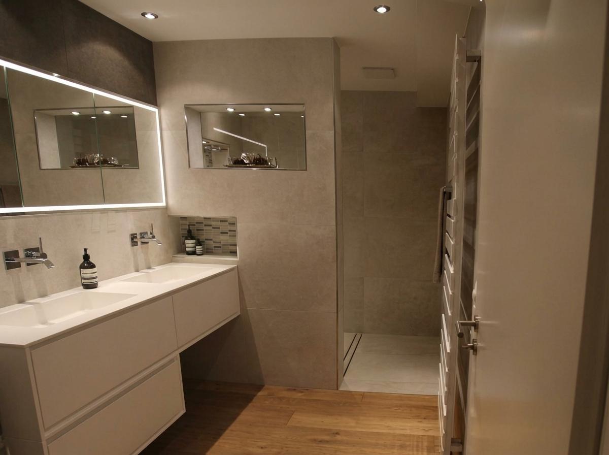 Modernes Badezimmer mit Doppelwaschtisch, zwei weißen Waschbecken und wandmontierten Chromarmaturen. Über dem Waschtisch befindet sich ein großer Spiegel mit umlaufender LED-Beleuchtung. Rechts davon ist eine Nische mit Spiegel und darunter eine weitere Nische mit Mosaikfliesen und Seifenspendern. Der Boden ist aus Holz. Im Hintergrund ist eine begehbare Dusche und ein weißer Handtuchwärmer mit einem Handtuch zu sehen.