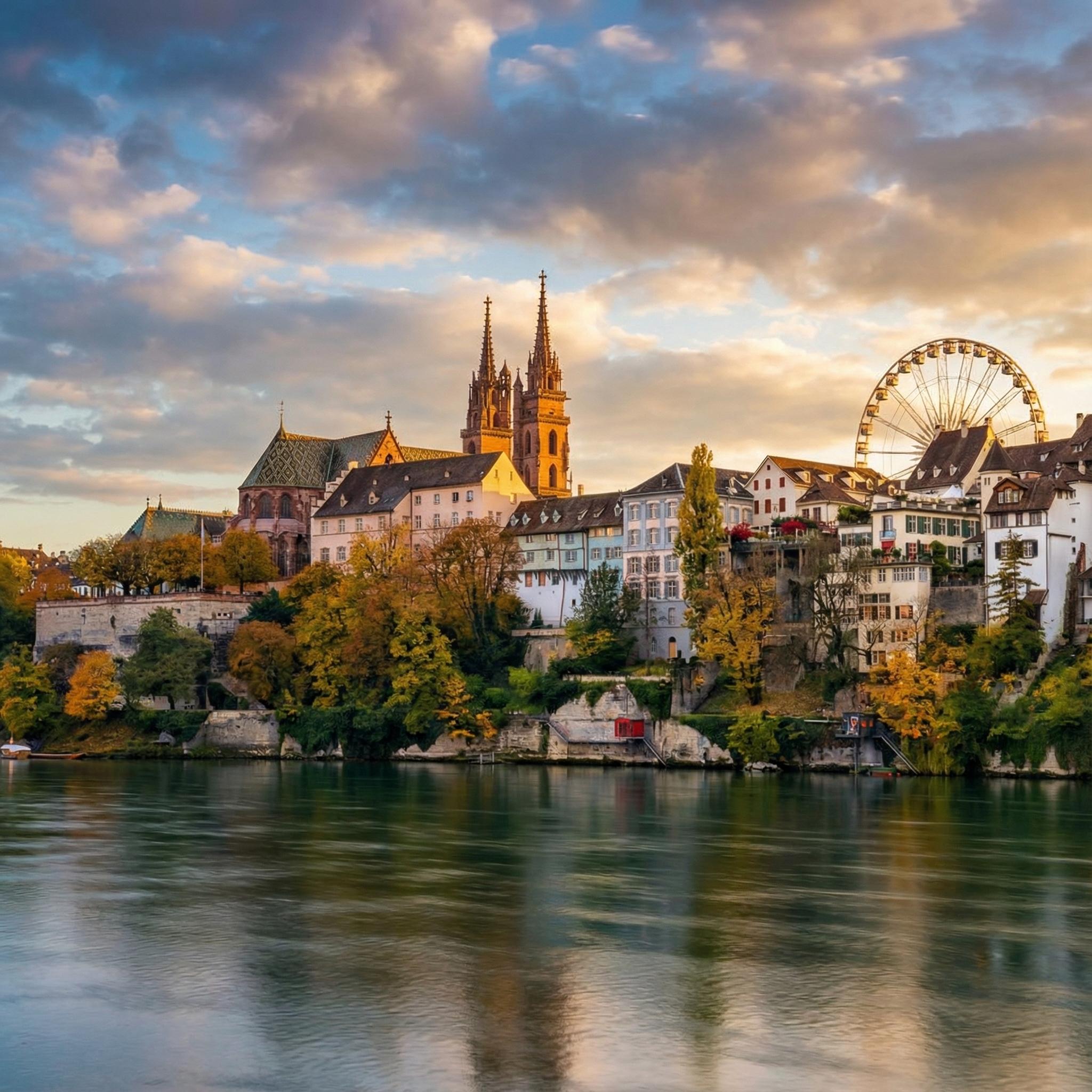Die historische Altstadt von Basel am Rhein bei Sonnenuntergang. Im Zentrum steht das Basler Münster mit seinen zwei Türmen und dem grünen Ziegeldach. Rechts ist ein großes Riesenrad zu sehen. Herbstliche Bäume säumen das Flussufer mit alten Gebäuden. Der Himmel zeigt blaue und orange Wolken, die sich im Wasser spiegeln.