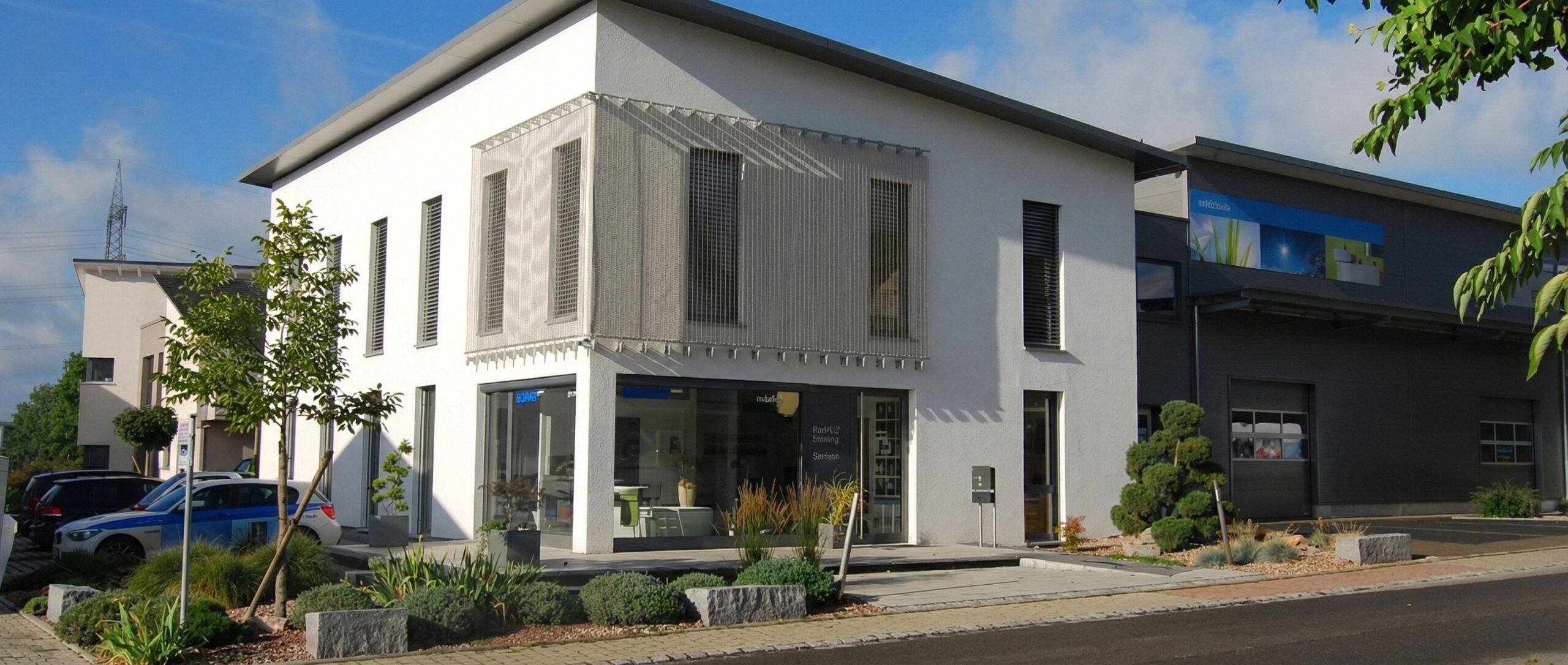 Modernes, weißes zweistöckiges Geschäftsgebäude mit großen Glasfenstern im Erdgeschoss, die Schaufenster mit Schriftzügen wie "Parkett", "Stalung" und "mister" zeigen. Die Obergeschossfenster sind mit externen, vertikalen Jalousien und einer dekorativen Metallgitterverkleidung versehen. Rechts daneben steht ein dunkelgraues Geschäftsgebäude mit zwei Garagentoren und einem großen blauen Werbebanner, auf dem "elektrotechnik" zu lesen ist. Die Gebäude sind von gepflegten Beeten mit Sträuchern, kleinen Bäumen und dekorativen Steinquadern umgeben, die an einen Gehweg und eine Straße grenzen. Im Hintergrund sind ein Strommast und ein weiterer Wohnkomplex unter einem klaren blauen Himmel sichtbar.